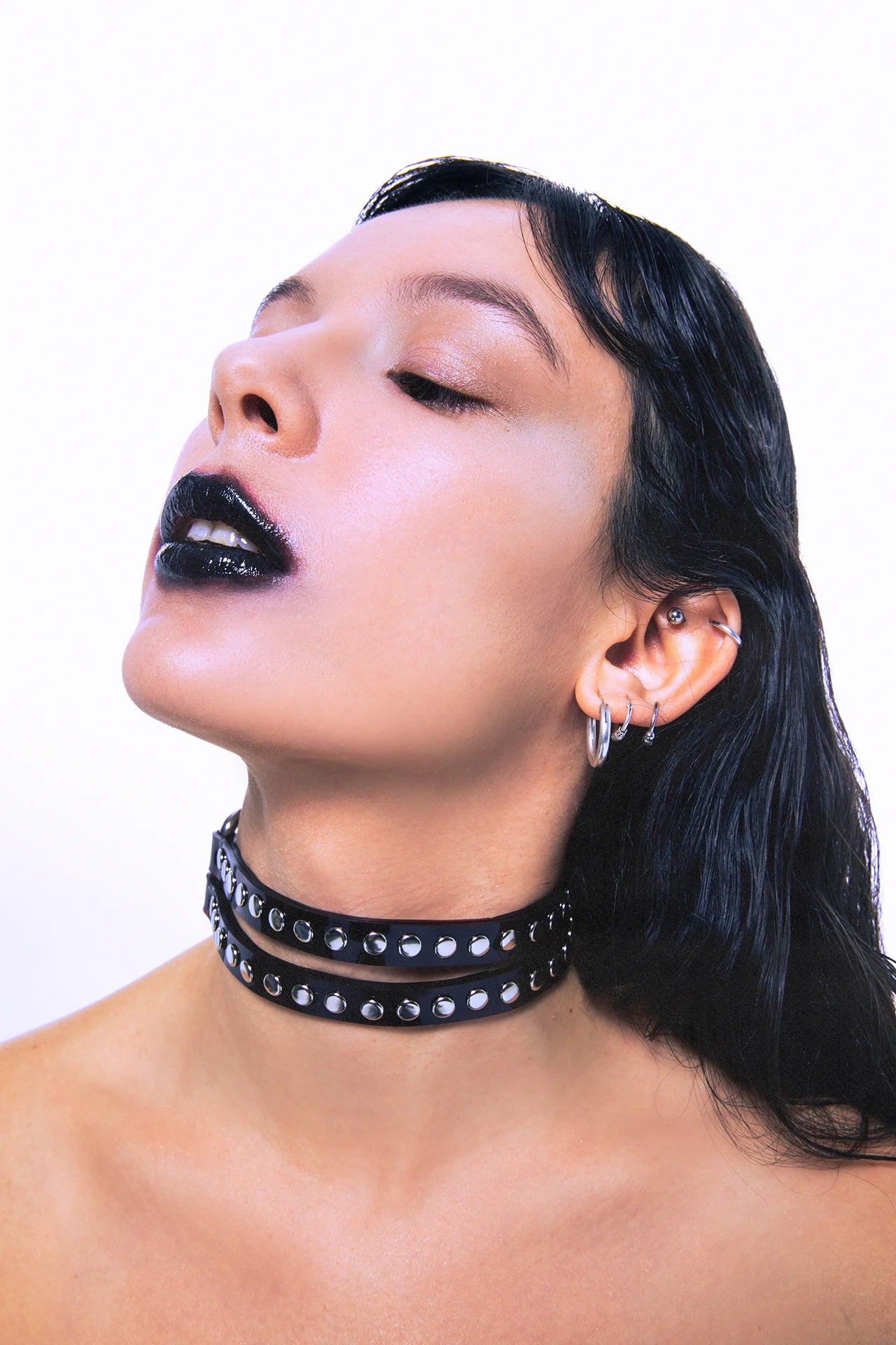 Side view of a glossy black double-layer PVC choker with silver rivets, worn by a model with wet-look hair and bold makeup – edgy unisex neckwear.