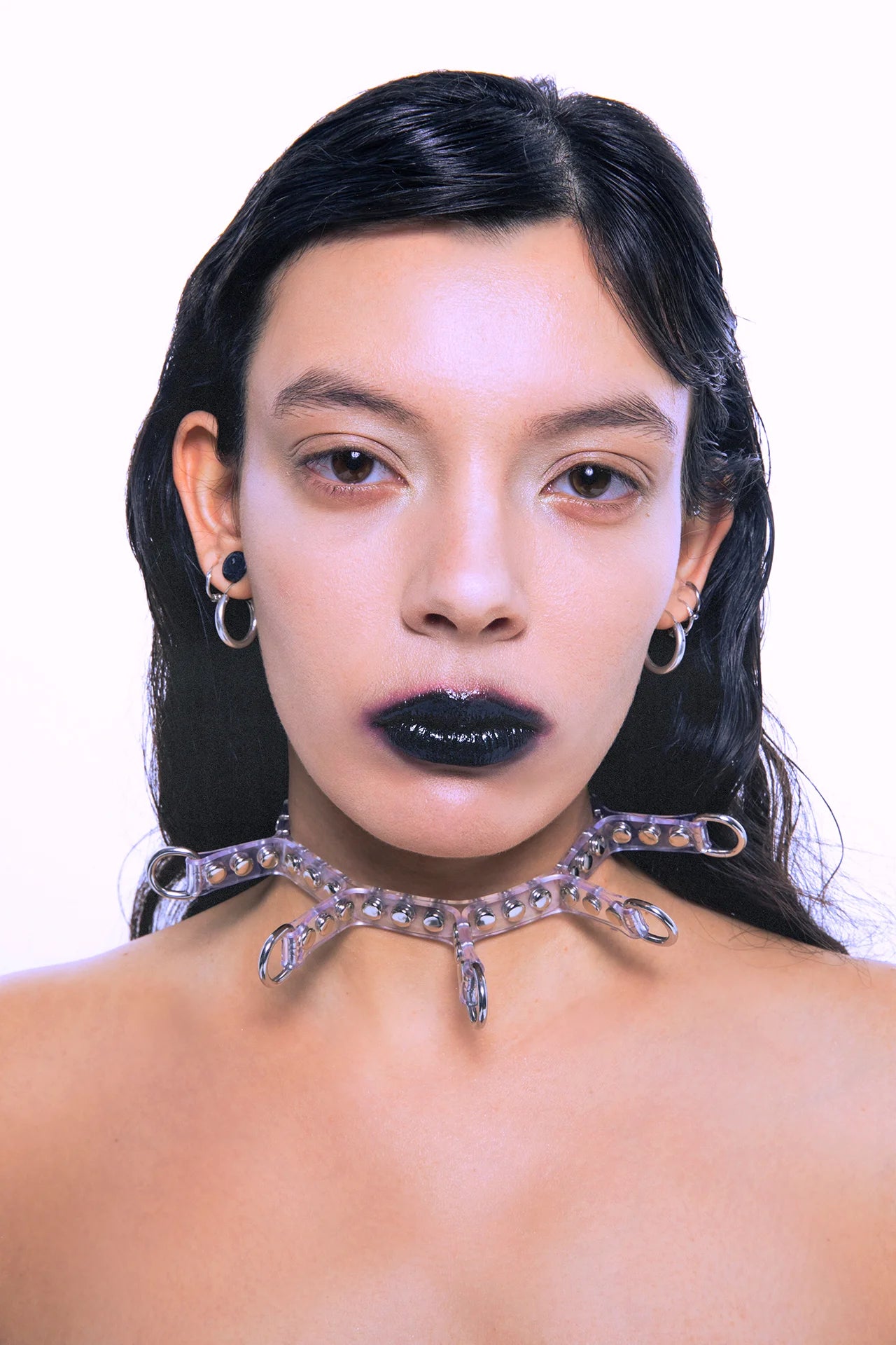 Close-up of a model wearing a transparent PVC choker with silver studs and multiple D-rings, styled with edgy black lipstick and silver earrings.