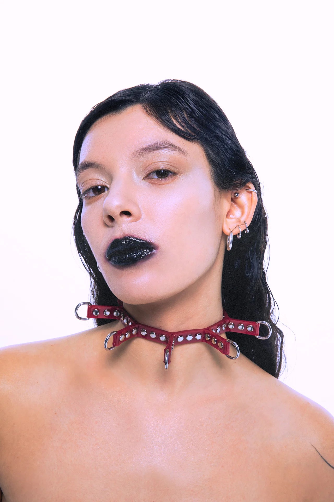 Model wearing a red spiked PVC choker with silver studs and D-rings, styled with wet-look hair and bold black lipstick for a goth-inspired look.