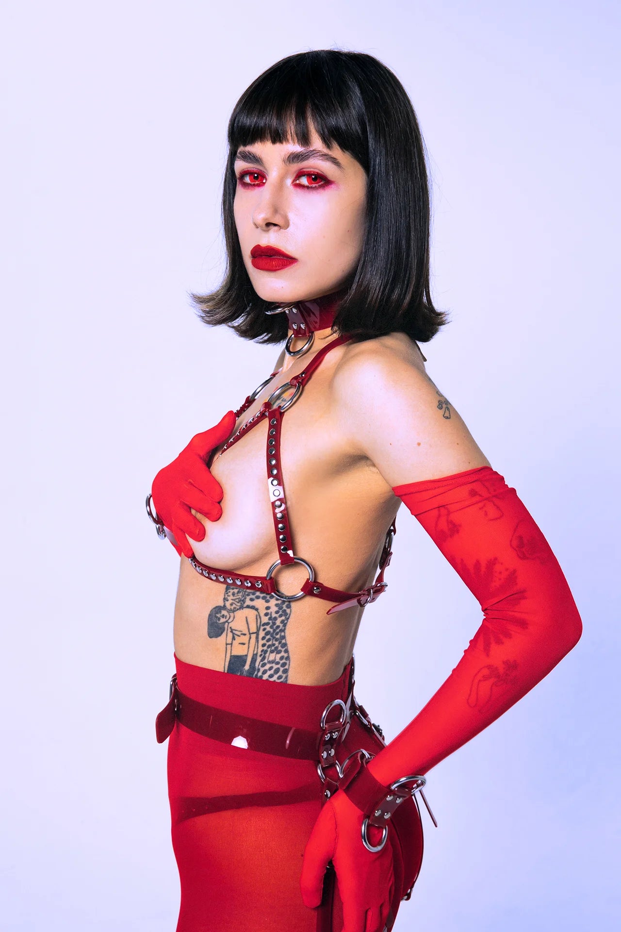 Side profile of a model in a red harness bra, transparent tights, red gloves, and a PVC belt. The styling is bold, gender-fluid, and designed for expressive fashion, ravewear, or provocative photoshoots.