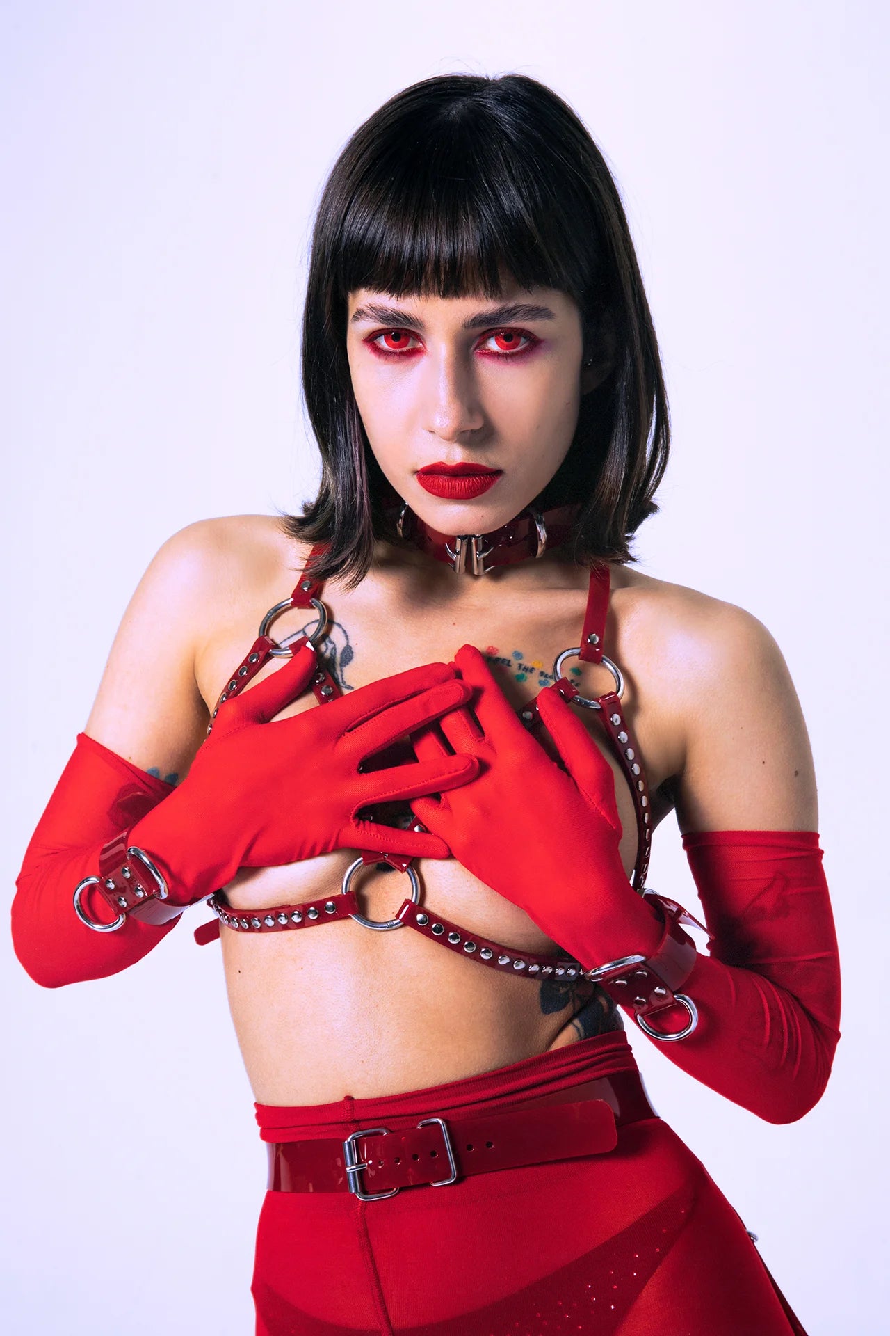 Close-up of a model wearing a red PVC harness bra, red gloves, and a matching choker collar. Silver hardware, bold red eye makeup, and glossy textures create a powerful alternative fashion statement for clubwear, kink, and queer aesthetics.