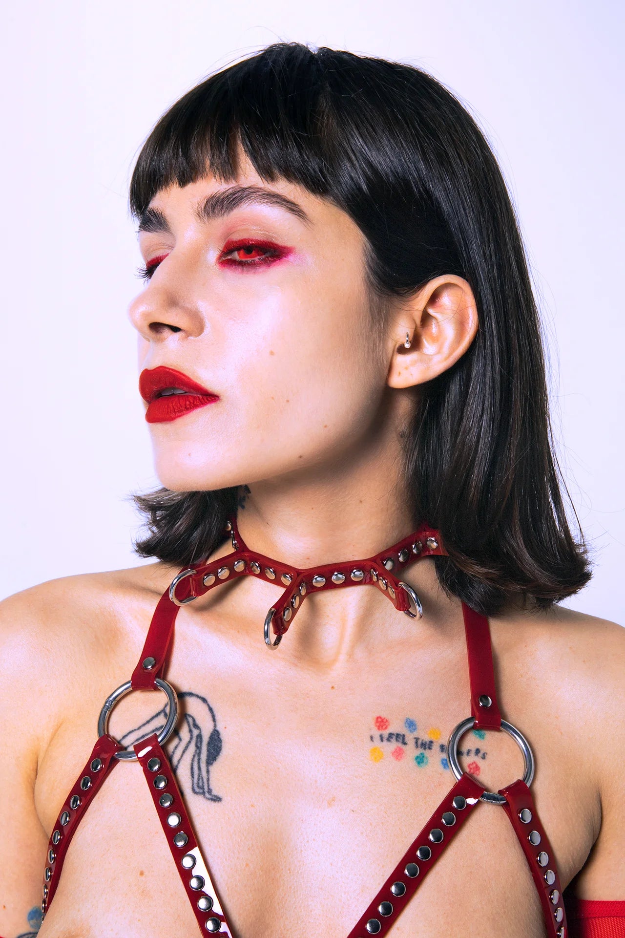 Model wearing a red vinyl spiked collar with silver D-rings, matched with red gloves and a studded harness bra, creating a bold fetishwear statement.