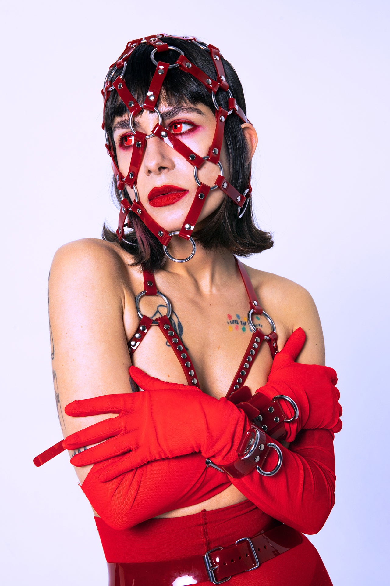 Model wears a red PVC bondage-style facial harness over a matching red bra with O-rings, rivets, and red gloves. The full-face cage harness creates a dramatic, avant-garde effect ideal for cyberpunk editorials and fetishwear fans.