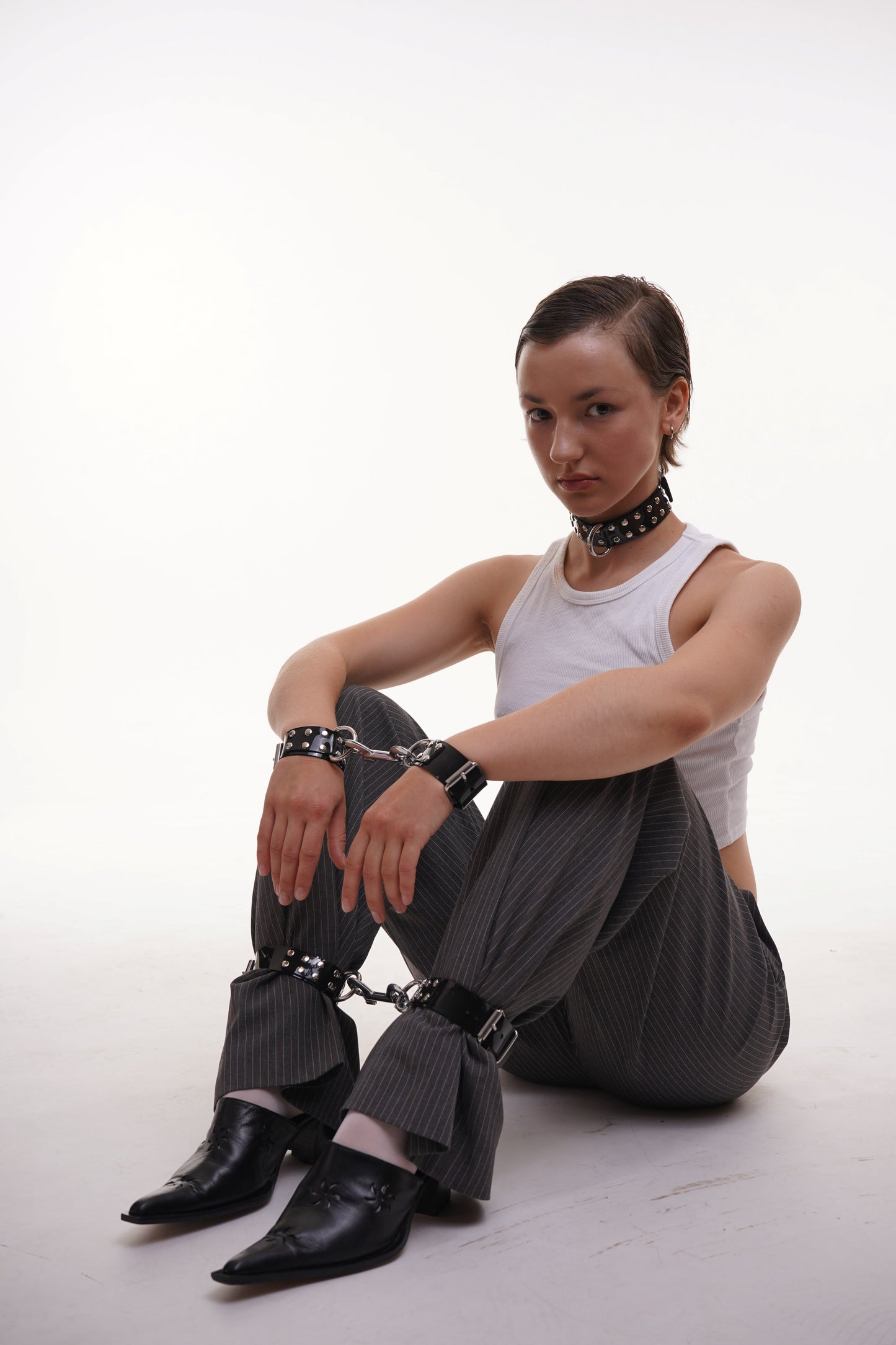 Model sitting on floor in a white tank top and pinstripe pants, wearing black PVC bondage cuffs connected with metal chains on wrists and ankles, styled with a studded choker – edgy alt fashion editorial.