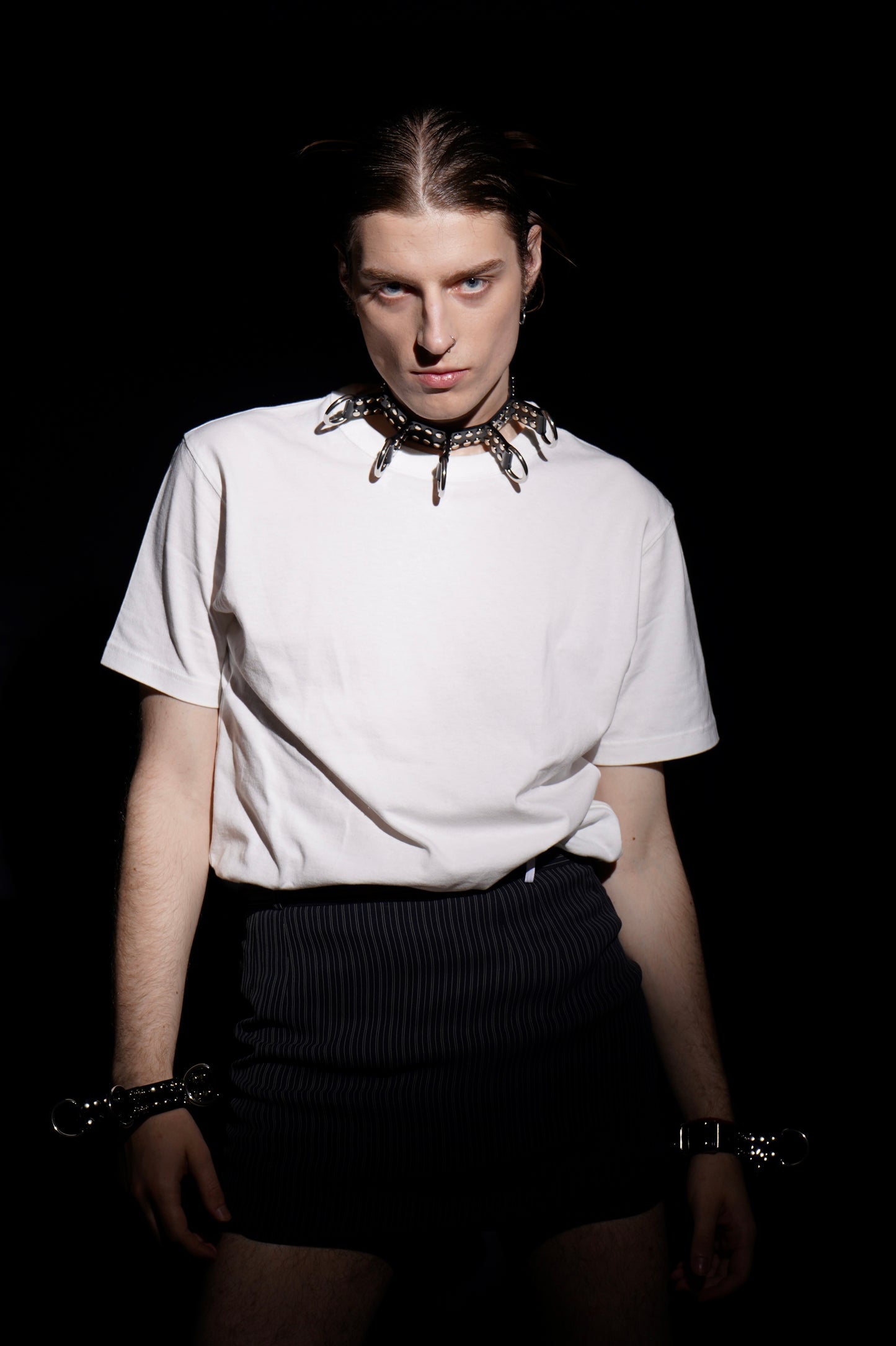 Model dressed in a plain white t-shirt and mini pinstripe skirt, spotlighted against a black background while wearing a spiked black PVC choker with silver rings. The look fuses minimalist wardrobe pieces with fetish elements, highlighting the versatility of this handmade cyberpunk accessory.