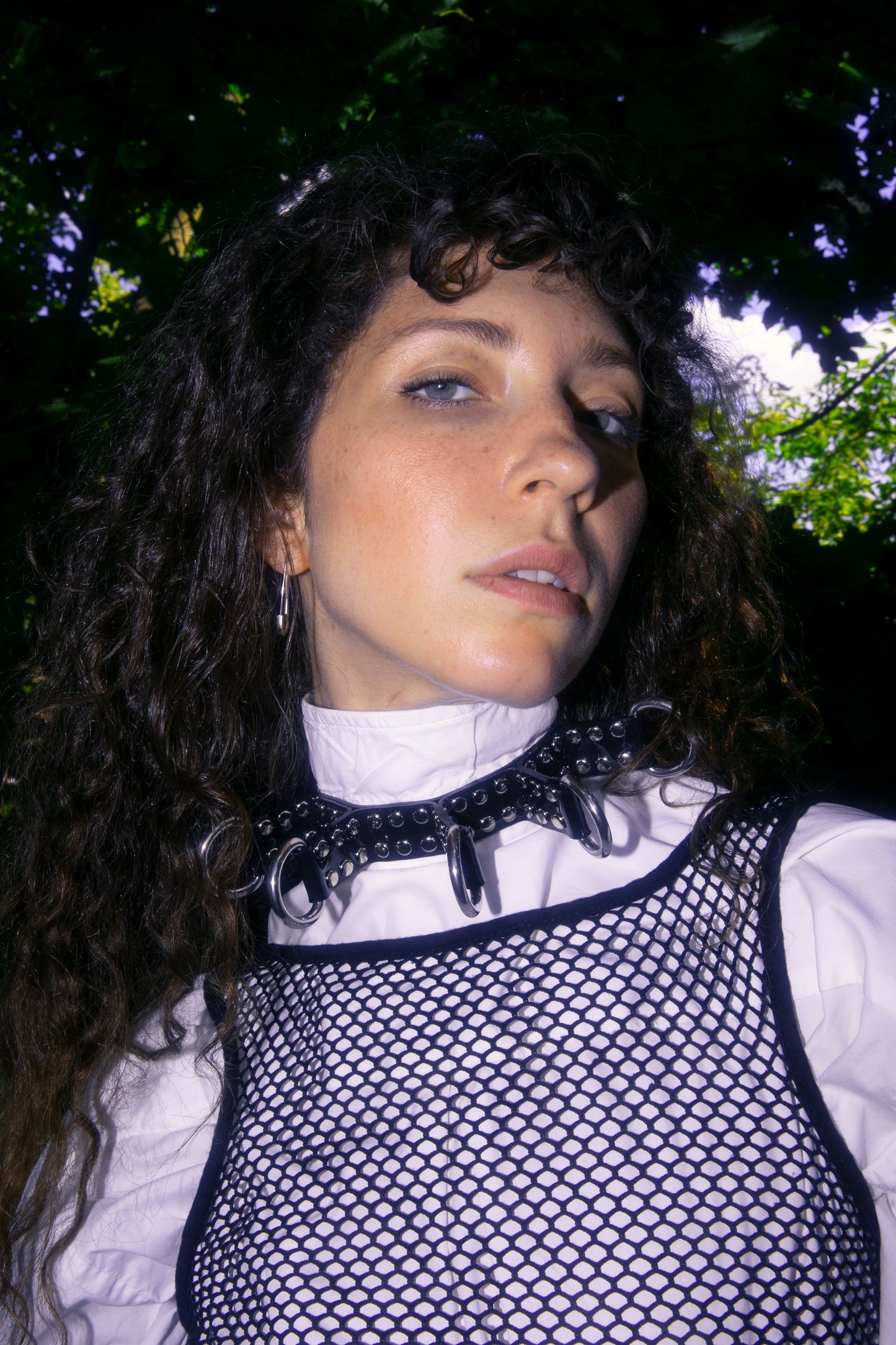 In a forest setting, a model gazes confidently at the camera wearing a black PVC choker with large D-rings layered over a white shirt and black mesh top. The edgy layering and nature contrast make this handcrafted bondage collar a statement for festival fashion, cyberpunk street style, or modern gothic aesthetics.