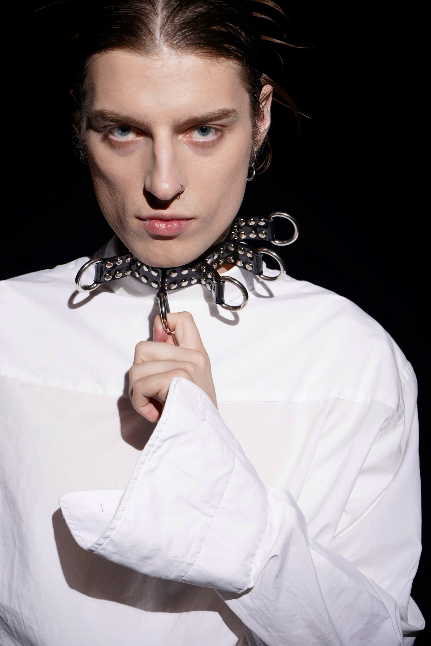 Close-up of a model wearing a black PVC bondage choker featuring silver eyelets, large O-rings, and spiked strap details. Styled with a crisp oversized white shirt, this unisex fetish accessory channels cyberpunk and BDSM fashion aesthetics. A bold, handcrafted alternative statement piece made in Berlin.