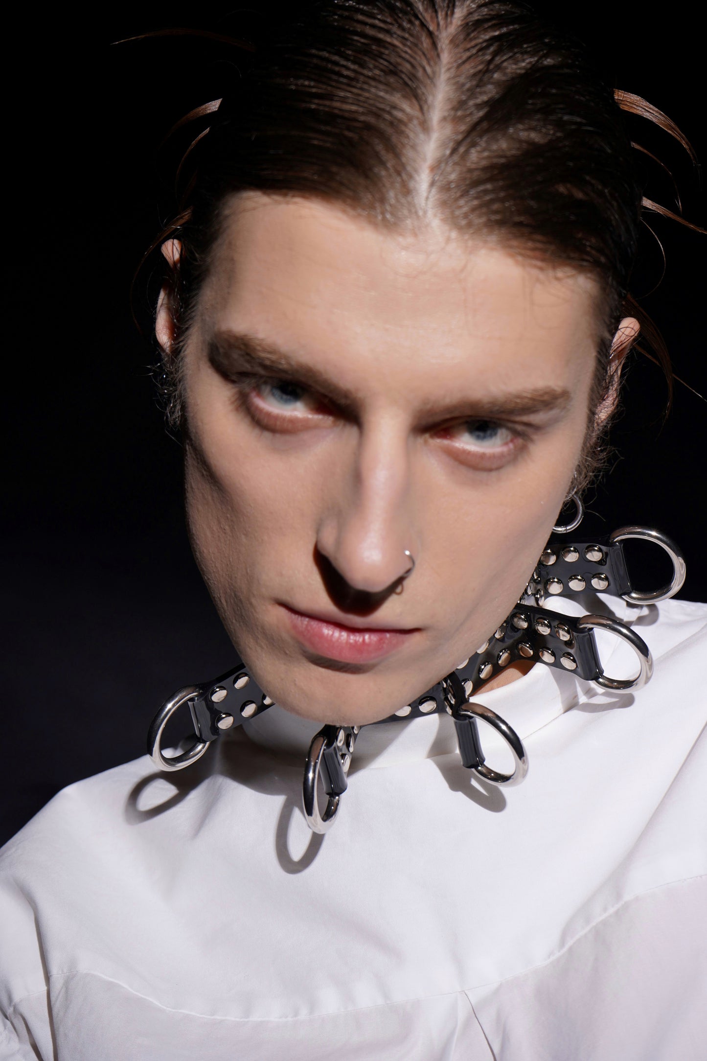 Close-up of a model wearing a black PVC bondage choker featuring silver eyelets, large O-rings, and spiked strap details. Styled with a crisp oversized white shirt, this unisex fetish accessory channels cyberpunk and BDSM fashion aesthetics. A bold, handcrafted alternative statement piece made in Berlin.