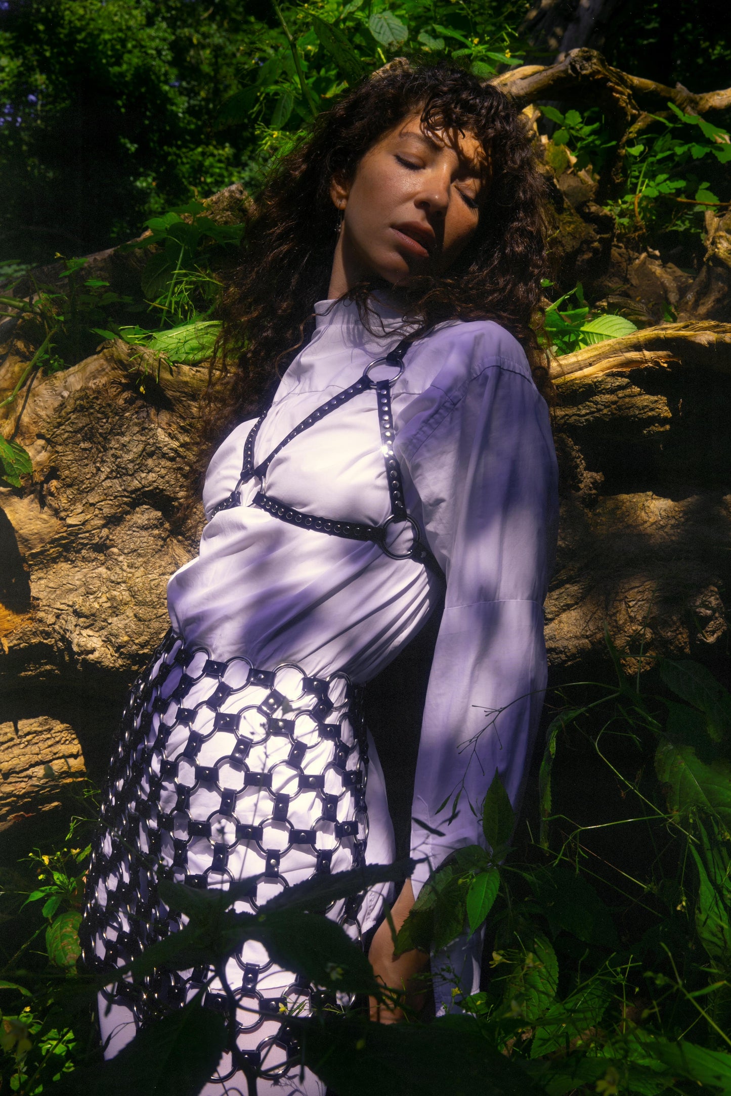 Model wearing a black PVC harness bra and geometric PVC skirt with hexagon-ring design, styled over a white shirt and photographed in a lush forest. This handcrafted fetishwear set blends nature with cyberpunk fashion, ideal for alternative styling, queer fashion editorials, and handmade harness looks from Berlin.