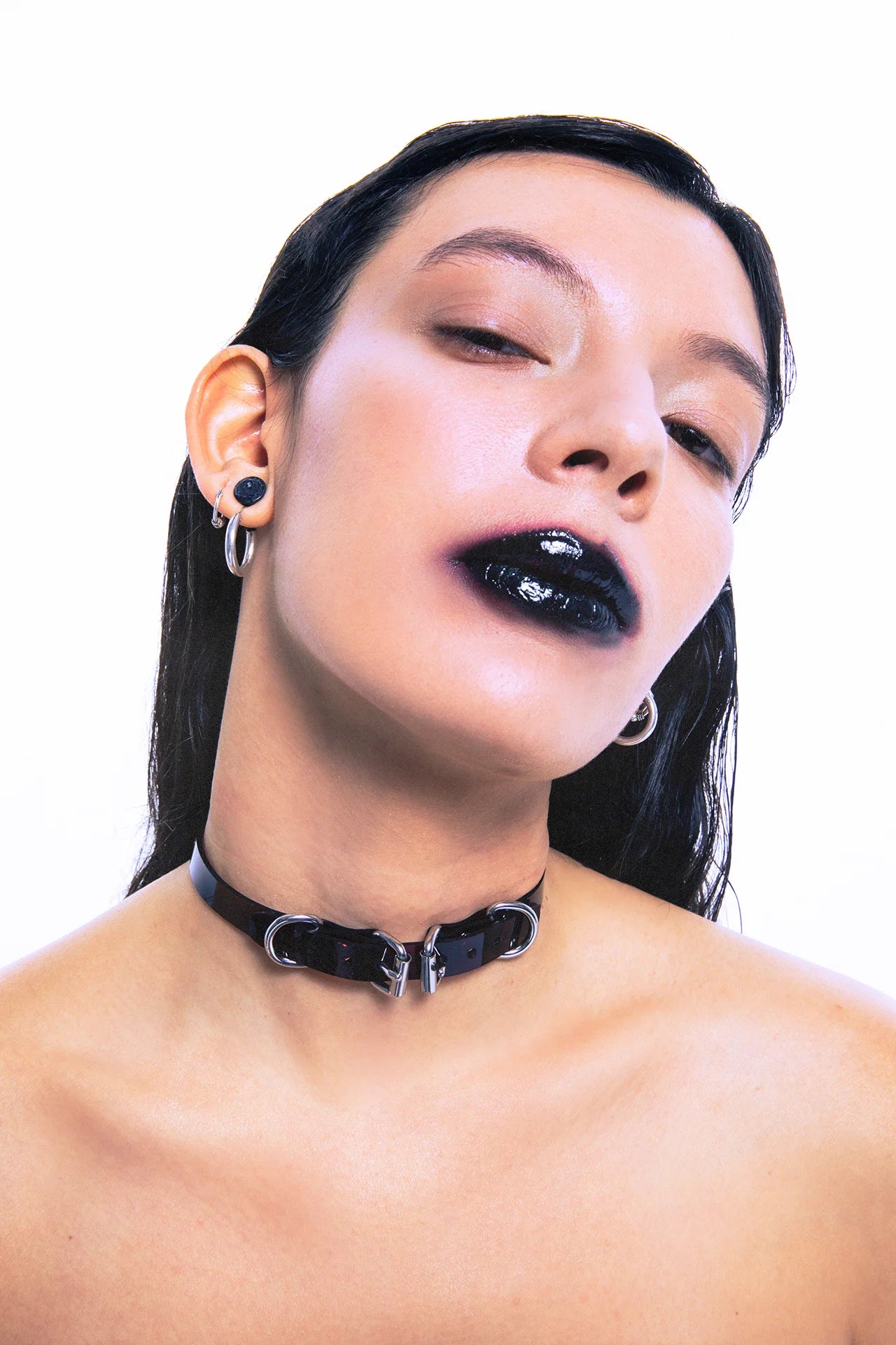 Close-up portrait of a model wearing a black patent PVC choker with dual D-rings and central padlock clasp, paired with glossy black lipstick and multiple silver ear piercings – edgy BDSM-inspired fashion accessory for alternative, fetish, and punk styles.