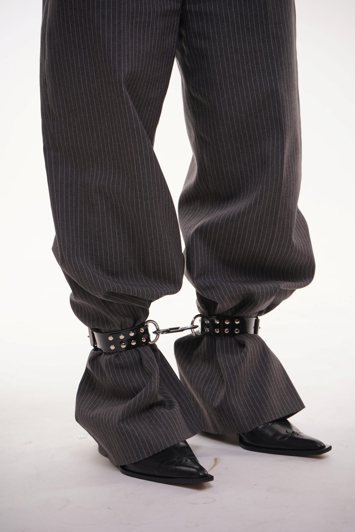 Focus on black patent ankle cuffs with silver studs and D-rings, fastened over wide pinstripe trousers – alternative styling for rave, goth or bondage wear.