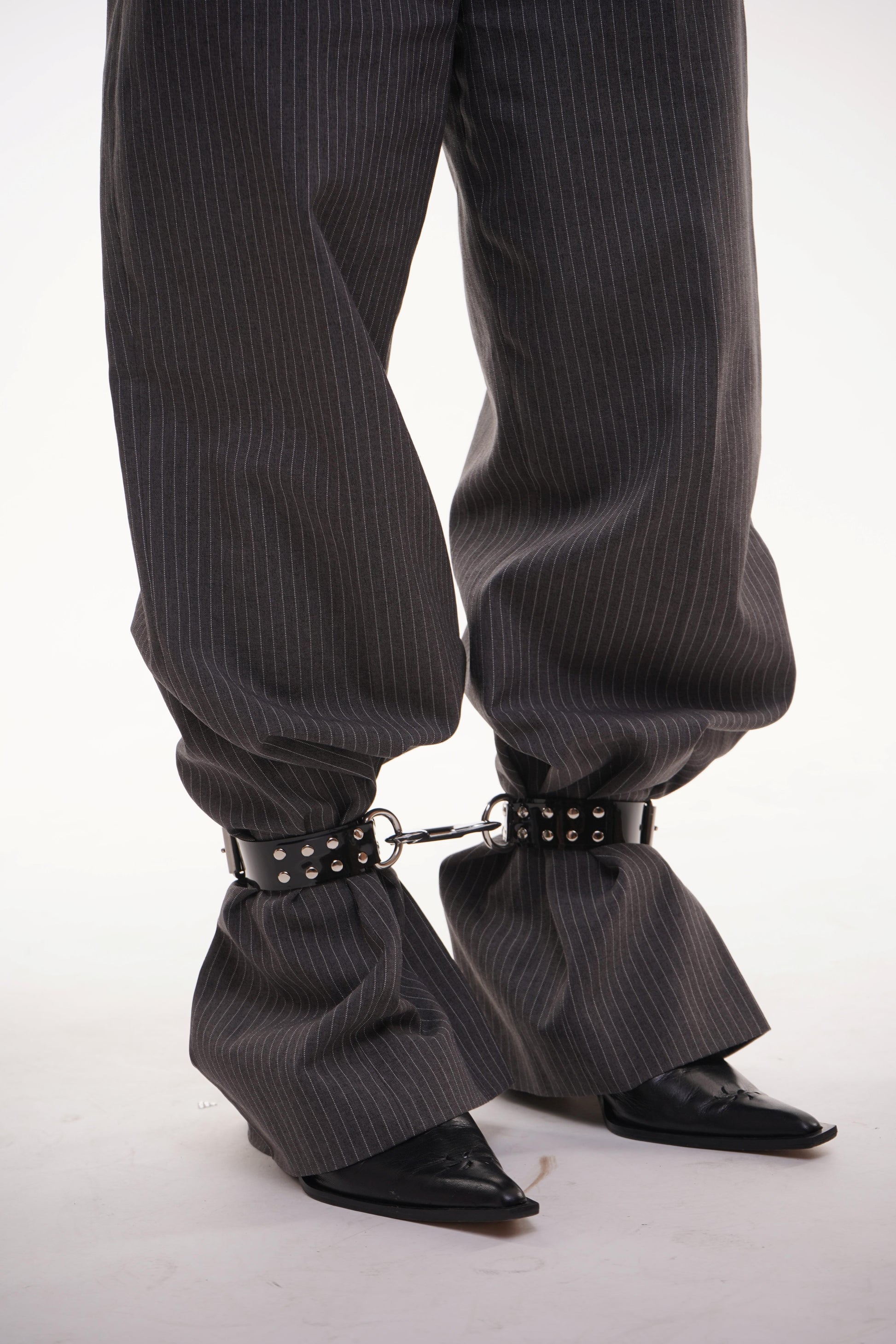 Focus on black patent ankle cuffs with silver studs and D-rings, fastened over wide pinstripe trousers – alternative styling for rave, goth or bondage wear.