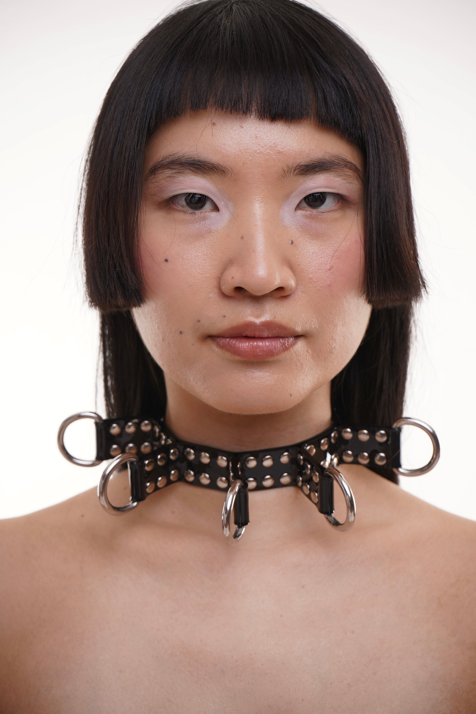 Model in front of a neutral backdrop wears a wide PVC BDSM collar featuring five silver O-rings and studded rivets. The hair is sleek and sharply cut, enhancing the futuristic feel of this handcrafted fetish fashion choker. Unisex and adjustable, it’s perfect for bold editorial styling or night-out looks.