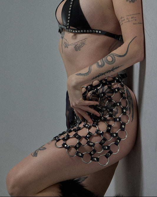 A model with red curly hair and tattoos lies on a gray studio floor wearing a black studded bra and a black mesh chain skirt.
