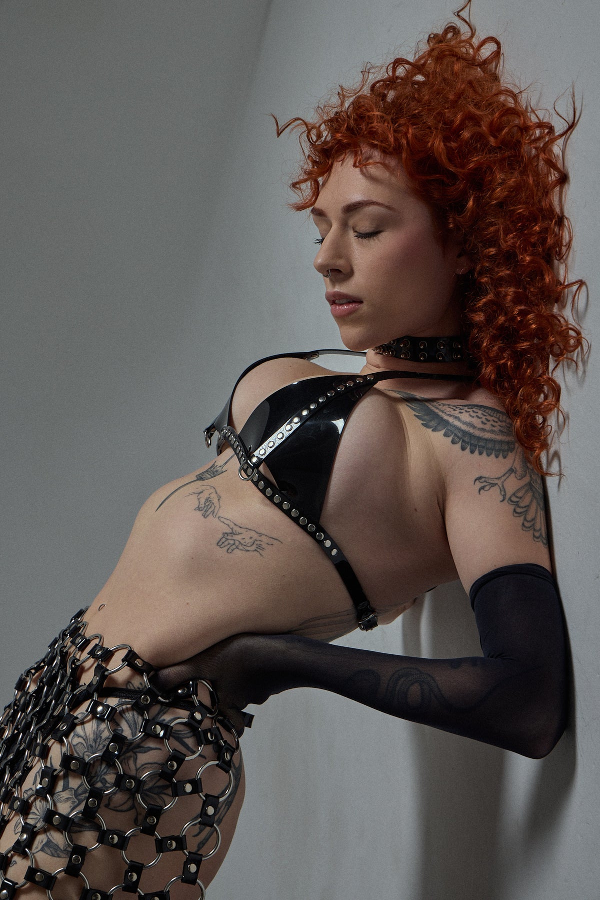 A model with vibrant red curly hair poses in a black studded strappy bra, a matching black studded choker, sheer black arm sleeves, and a black studded fishnet skirt against a plain gray wall.